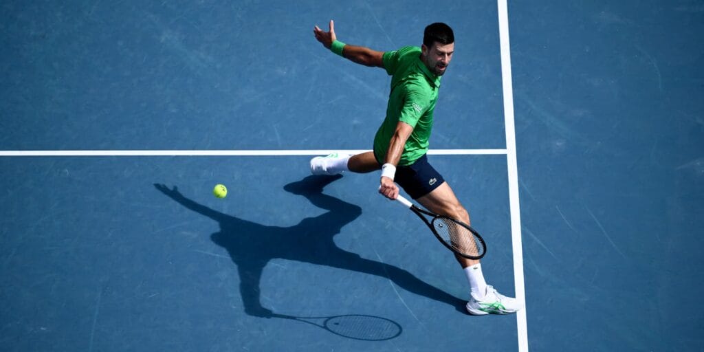 Novak Djokovic hitting a ball at the Australian Open wearing a green shirt and blue shorts